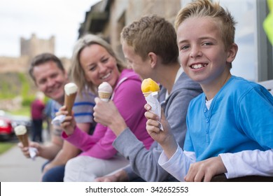 Family enjoying ice cream outside of a shop in a village. They are all holding their cones and a castle can be seen in the background in the distance. 