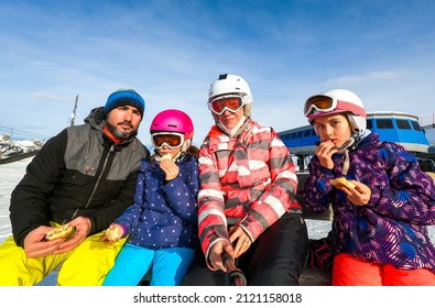 Family Enjoying Lunch On A Break On Vacations In Skiing Gear. Family With Children On Skiing Vacation Eating Sandwiches In Helmets And Goggles. Winter Sports Mountain Resort.