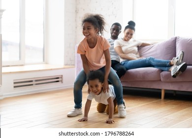 Family evening at home. Happy loving african married couple embracing on sofa in living room watching their two kids daughter and son enjoying funny active game, jumping and playing leapfrog on floor