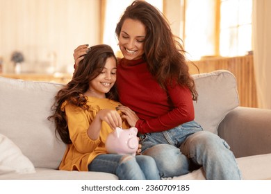 Family Finance Lessons. Arabic Mommy and Daughter Sharing Joy of Saving, Putting Money In Piggybank, Investing in a Kid's Financial Future, Sitting On Couch In Modern Living Room Indoors
