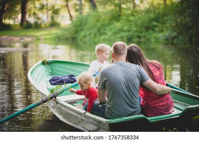 The family is floating on the lake in a boat with oars in the park, dad hugs mom, the girl looks into the water and holds a twig.