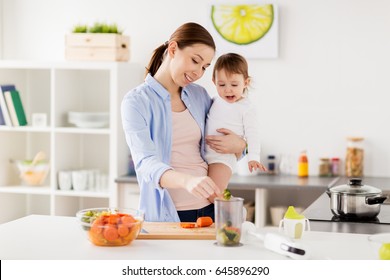 family, food, healthy eating, cooking and people concept - happy mother adding chopped vegetables to blender cup and holding little baby girl at home kitchen - Powered by Shutterstock