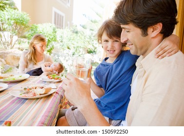 Family Of Four Gathering Around A Table With Food In A Home Porch Garden Outdoors With Mother, Father, Baby Girl, And A Young Boy Drinking Water With Dad.