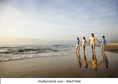 Family Of Four Holding Hands And Walking On Beach In North Carolina.