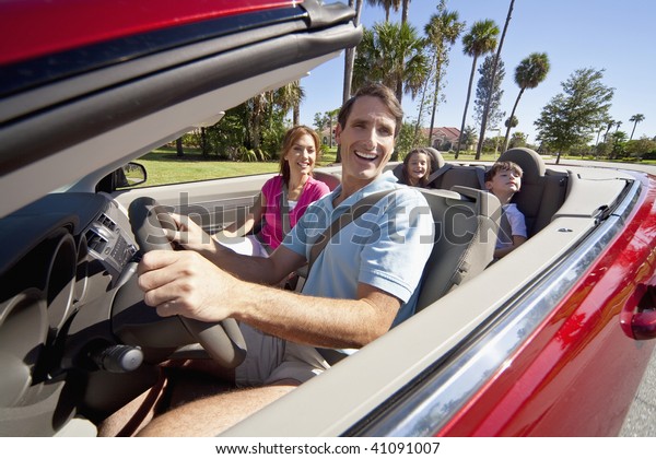 A family of four, mother, father, son and daughter\
driving in a convertible car on a sunny day in hot location with\
palm trees