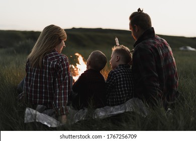 A Family Of Four People, Dad, Mom And Two Sons Are Sitting Around The Fire. Warm Marshmallows And Enjoy Outdoor Recreation. Vacation Concept In The Country. Tourism In The Summer.