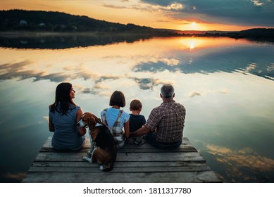 family of four people sitting on the shore on a wooden bridge of a large lake in summer and watching the beautiful sunset with dog back view - Powered by Shutterstock