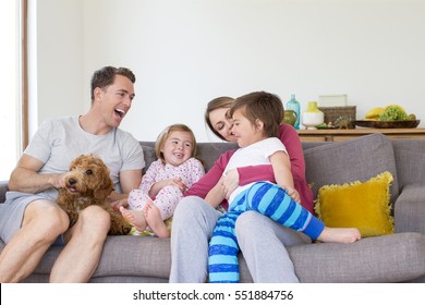 Family Of Four Are Sitting On The Sofa In The Living Room Of Their Home Together With Their Pet Dog. 