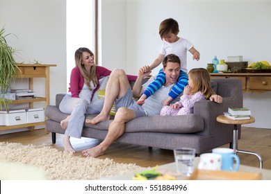 Family Of Four Are Sitting On The Sofa In The Living Room Of Their Home Together. The Father Has His Son On His Shoulders And The Little Girl Is Sitting Next To Him. 