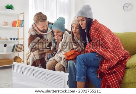 Family freezing at home. Sad mom, dad and children wearing warm plaids and winter hats sitting on sofa by portable electric heater inside very cold residential flat