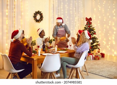 Family and friends dining at home celebrating Christmas eve with traditional food and decoration. Multiracial people sit at table in living room and applaud their male friend who serves delicious meal
