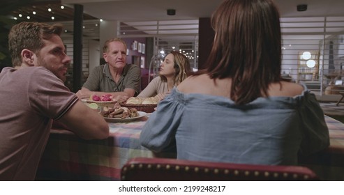 Family And Friends Gathered Outside The House At Night In Home Backyard. Group Of People Converse Sitting At Dinner Table.