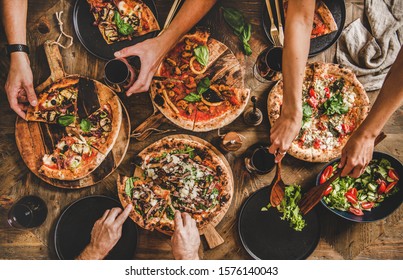 Family Or Friends Having Pizza Party Dinner. Flat-lay Of People Cutting And Eating Italian Pizza And Drinking Red Wine From Glasses Over Wooden Table, Top View. Fast Food Lunch, Gathering, Celebration