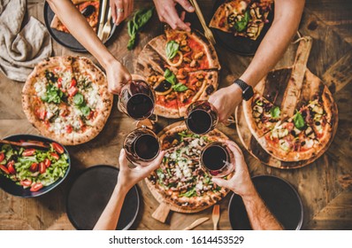 Family Or Friends Having Pizza Party Dinner. Flat-lay Of People Clinking Glasses With Red Wine Over Rustic Wooden Table With Various Kinds Of Italian Pizza, Top View. Fast Food Lunch, Celebration