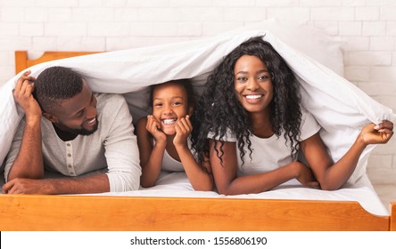 Family fun. Cheerful black mother, father and daughter hiding under blanket, having fun together at home, panorama - Powered by Shutterstock