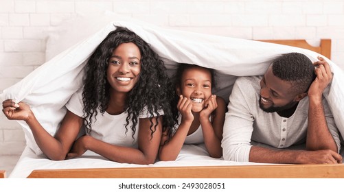 Family fun. Cheerful black mother, father and daughter hiding under blanket, having fun together at home, panorama - Powered by Shutterstock