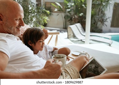 Family Fun At Home. Dad And Little Boy Sitting On Sofa In Tropical Villa Patio With Tablet. Father Drinking Tea And Using Portable Digital Device. Enjoying Leisure On Weekend At Resort.