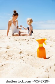 Family Fun On White Sand. Closeup On Sunscreen Bottle At Sandy Beach On A Sunny Day. Mother And Child In Swimsuits Playing In Background