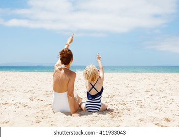 Family Fun On White Sand. Seen From Behind Mother And Child In Swimsuits Sitting At Sandy Beach On A Sunny Day And Pointing Up On Something