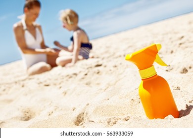 Family Fun On White Sand. Closeup On Sunscreen Bottle At Sandy Beach On A Sunny Day. Mother And Child In Swimsuits Playing In Background
