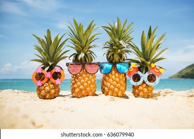 Family Of Funny Attractive Pineapples In Stylish Sunglasses On The Sand Against Turquoise Sea. Tropical Summer Vacation Concept. Happy Sunny Day On The Beach Of Tropical Island. Family Holiday