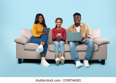 Family Gadgets. Excited Young Black Parents And Their Daughter Using Different Electronic Devices While Sitting On Couch Over Blue Studio Background