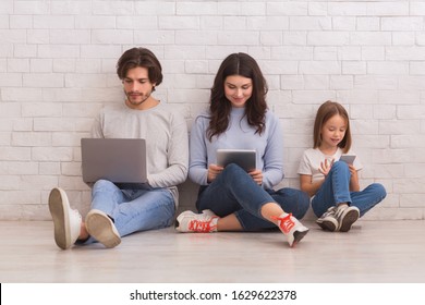 Family Gadgets. Happy Parents And Their Little Daughter Holding Different Electronic Devices In Hands While Sitting On Floor Near White Brick Wall