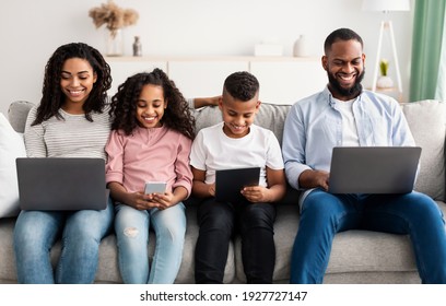 Family Gadgets. Portrait Of Happy African American Parents And Their Little Children Holding And Using Different Electronic Devices While Sitting On The Couch In Living Room At Home