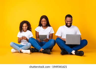 Family Gadgets. Portrait of happy African American parents and their little daughter holding and using different electronic devices while sitting on the floor over yellow studio background - Powered by Shutterstock