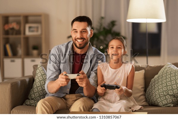 family, gaming\
and entertainment concept - happy father and little daughter with\
gamepads playing video game at\
home