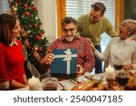 Family is gathered around a festive table enjoying their christmas dinner and giving a present to their father and grandfather