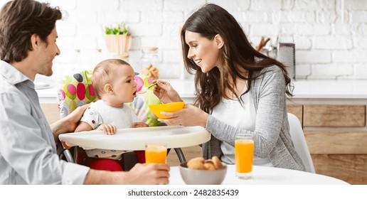 A family is gathered around a table in their kitchen, enjoying a meal together. A mother is feeding her baby from a bowl with a green spoon, while the father looks on, smiling - Powered by Shutterstock