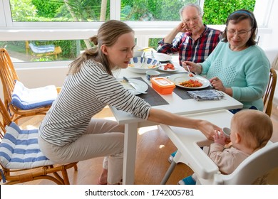 Family Gathering For Lunch At Home. Mom Teaching Baby Daughter Drink From Cup. Grandma And Grandpa Looking At Cute Granddaughter In High Baby Chair. Togetherness Or Child Care Concept