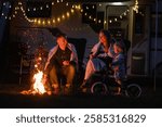 Family gathers around a warm campfire at their RV campsite, wrapped in blankets, enjoying drinks and sharing joyful moments with their child under twinkling string lights.