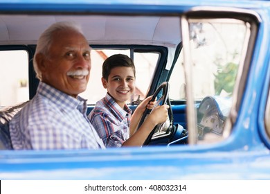 Family and Generation gap. Old grandpa spending time with his grandson. He teaches him to drive. The boy holds the volante of a vintage car from the 60s. They both smile happy looking at camera.