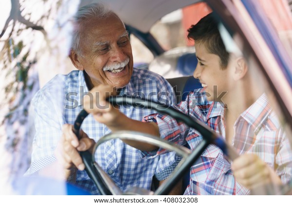Family and Generation gap. Old grandpa spending\
time with his grandson and teaching him to drive. The boy holds the\
volante of a vintage car from the 60s. They both smile happy\
looking each other.