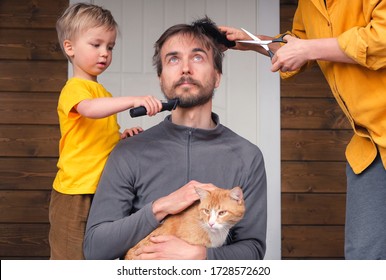 Family Haircut At Home During Quarantine Lockdown, Domestic Hairdressers. Mother Cutting Hair To Father And Little Child Boy Cut Dad Beard With Clipper. Beauty And Selfcare At Home Lifestyle.