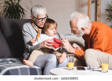 Family, Happiness, Generation, Home And People Concept - Happy Grandparents Sitting On Sofa With One Year Old Grandchild. Giving Birthday Present