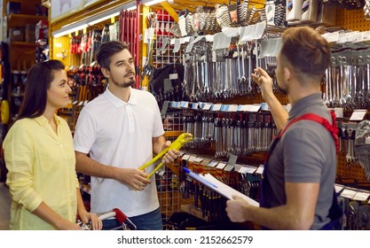 Family in hardware store. Seller advises couple of customers on type of spanners in building materials store. Husband and wife buy tools for repairs, listen carefully to employee of store in overalls. - Shutterstock ID 2152662579