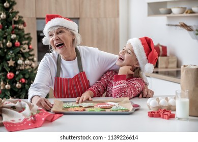 Family Harmony. Portrait Of Happy Grandmother And Granddaughter Laughing While Standing In Kitchen. Tray The Christmas Cookies On Table