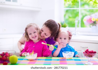 Family Having Breakfast In A White Sunny Kitchen. Young Mother Feeding Two Kids, Eating Fruit And Dairy. Healthy Nutrition For Children. Parent With Toddler Kid And Baby Cooking Morning Meal.