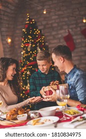 Family Having Christmas Dinner At Home, Gathered Around The Table, Enjoying Their Time Together; Mother And Father Giving Christmas Present To Their Son