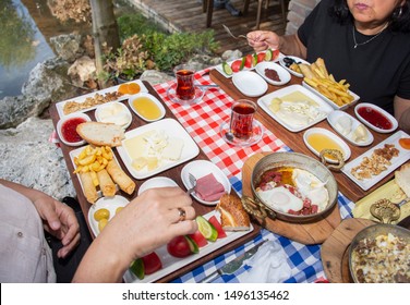 Family Having Delicious Rich Traditional Turkish Breakfast Include Tomatoes, Cucumbers, Cheese, Butter, Eggs, Honey, Bread, Olives, Jam, Sausage, Fried Potatoes And Tea Cups. 