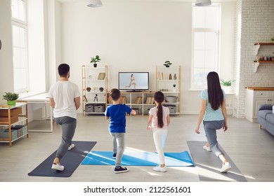 Family Having Fitness Workout At Home. Back View Of Happy Mum, Dad And Kids Standing On Rubber Mats Together, Watching Video Workout With Gym Instructor On TV Screen And Doing Leg Stretching Exercise