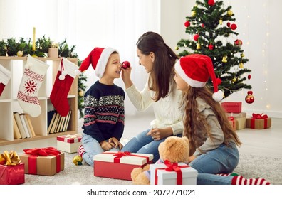 Family having fun and enjoying good time together on Christmas holidays at home. Happy mom and two children in red Santa caps playing with Xmas tree baubles sitting on floor in cozy, decorated house - Powered by Shutterstock