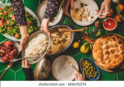 Family having Turkish dinner. Flat-lay of people passing rice pilav over green table with lamb in yogurt sauce, fresh arugula and strawberry salad and flatbread, top view. Ramazan iftar supper