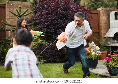 Family Are Having A Water Fight Together With Water Pistols In The Garden. The Little Girl Is Aiming For Her Dad, Who Is Aiming For The Little Boy. 