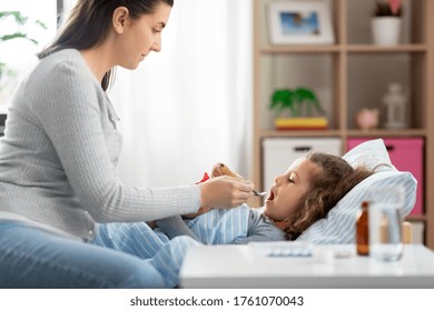 Family, Health And Medicine Concept - Mother Giving Cough Syrup To Little Sick Daughter Lying In Bed At Home