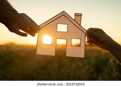 Family Holds Paper House At Sunset, Sun Shines Through Window. Hand Holding Paper Cut Of House Symbols At Sunset 