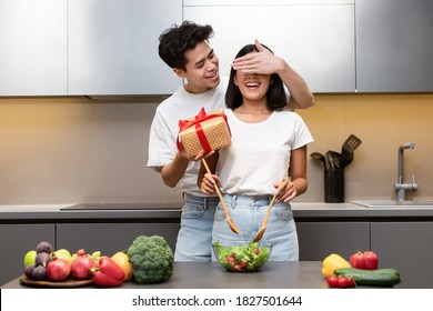 Family Holiday Celebration. Asian Husband Preparing Surprise Gift For Happy Wife Standing In Kitchen At Home. Couple Celebrating Birthday And Cooking Romantic Celebratory Dinner